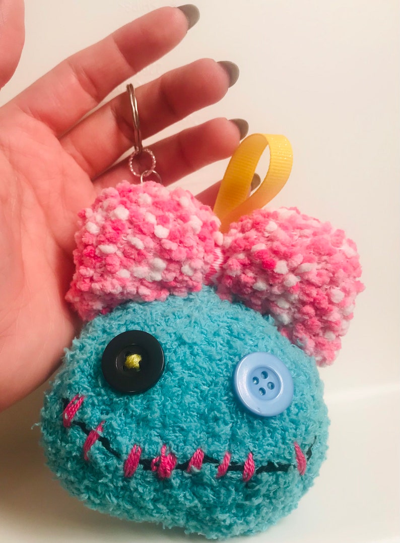 Pu&ograve; includere: Portachiavi in ​​peluche fatto a mano a forma di personaggio dei cartoni animati. Il personaggio ha un corpo blu con cuciture rosa, un fiocco rosa e bianco e due occhi a bottone. Sono attaccati un nastro giallo e un portachiavi argentato.