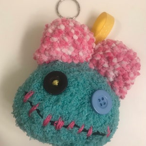 Pu&ograve; includere: Un portachiavi in ​​peluche fatto a mano a forma di personaggio dei cartoni animati. Il personaggio &egrave; turchese con orecchie a pompon rosa e bianche. Ha una bocca cucita rosa, occhi di bottoni neri e blu e un anello di nastro giallo.