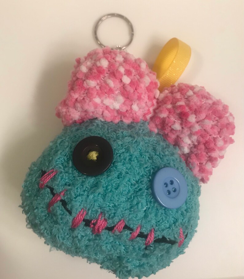 Pu&ograve; includere: Un portachiavi in ​​peluche fatto a mano a forma di personaggio dei cartoni animati. Il personaggio &egrave; turchese con orecchie a pompon rosa e bianche. Ha una bocca cucita rosa, occhi di bottoni neri e blu e un anello di nastro giallo.