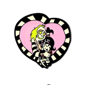 May include: Enamel pin featuring a cartoon illustration of two characters embracing within a pink heart. The heart is framed by a black and white striped border. One character has green hair and the other has black hair.