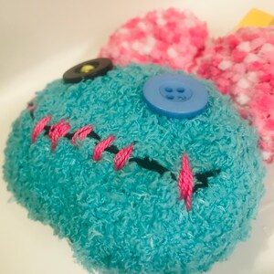Pu&ograve; includere: Un giocattolo di peluche fatto a mano con un corpo turchese, bocca cucita in nero e dettagli in filo rosa. Presenta un bottone blu e un bottone nero per gli occhi e orecchie soffici rosa e bianche.