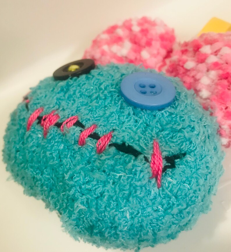 Pu&ograve; includere: Un giocattolo di peluche fatto a mano con un corpo turchese, bocca cucita in nero e dettagli in filo rosa. Presenta un bottone blu e un bottone nero per gli occhi e orecchie soffici rosa e bianche.