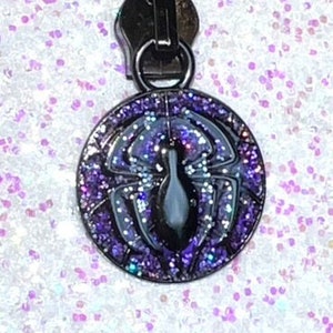 May include: Black zipper pull with a purple and black spider web design. The spider is in the center of the web.