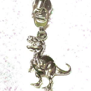 May include: A silver-toned zipper pull featuring a detailed Tyrannosaurus Rex dinosaur charm. The charm is attached to the zipper pull with a small ring. The background is a soft, out-of-focus white with subtle pink and green accents.