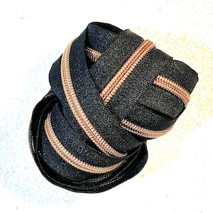 Puede incluir: Cremallera negra enrollada con una cinta de cremallera de color cobre metálico. La cremallera está enrollada firmemente, mostrando el contraste entre la tela oscura y los dientes metálicos brillantes. Para proyectos de costura.
