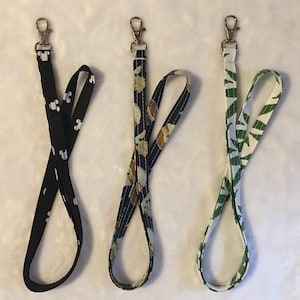 May include: Three fabric lanyards with different patterns. The first lanyard is black with a white Mickey Mouse pattern. The second lanyard is blue with a white and green pattern of a baby Yoda character. The third lanyard is white with a green and brown pattern of leaves.