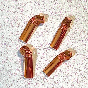 May include: Four rose gold zipper pulls arranged on a glittery white surface. Each pull is a rectangular shape with a curved top and a small hole for the zipper. The pulls are shiny and reflect light.