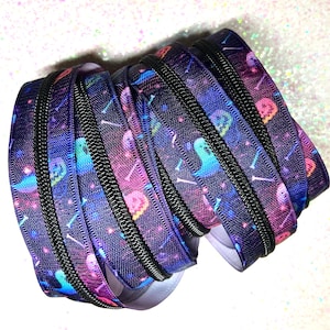 May include: A roll of black zipper tape with a colorful Halloween-themed print. The fabric features ghosts, skulls, and bones against a dark blue and purple galaxy background. The zipper teeth are black.