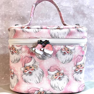 May include: A pink cosmetic bag with a white zipper and a handle. The bag is covered in a pattern of Santa Claus faces with snowflakes. The bag has a pink bow with a rhinestone embellishment.
