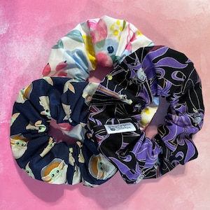 Puede incluir: Tres scrunchies de tela para el pelo. Uno es floral con flores rosas, blancas y amarillas. Otro es negro con un diseño morado y blanco con un personaje de una película de Disney. El último es azul marino con un diseño blanco y gris con un personaje de una serie de televisión popular. Los scrunchies tienen una pequeña etiqueta blanca que dice "Raggedie Stitchin".