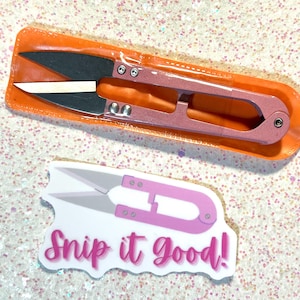 May include: Pink thread snips with black blades in an orange plastic package. A sticker with a pink thread snip graphic and the text "Snip it Good!" is also visible. The tools are likely for crafting or sewing.