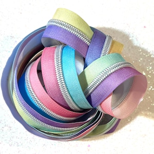 May include: A collection of colorful zipper tapes in shades of pink, purple, blue, and green. The tapes are arranged in a circular pattern and feature a silver zipper teeth.