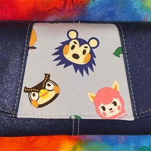 May include: A blue fabric wallet with a flap closure. The front of the wallet features a light blue fabric panel with a repeating pattern of animal characters from the Animal Crossing video game series.