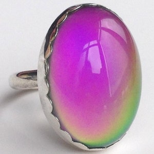 May include: A silver ring with a large oval-shaped mood ring stone that changes color. The stone is currently a vibrant purple with hints of green and yellow.