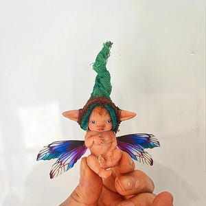 May include: A small, detailed fairy figurine with a green and brown hat, large ears, and colorful wings. The fairy is held in a hand against a white background. The wings have blue and purple hues.