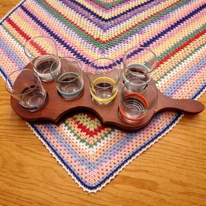May include: A wooden shot glass holder with six clear glasses, each with a colored band. The holder sits on a colorful, crocheted placemat. The glasses have bands of white, teal, yellow, and orange. The placemat has a rainbow pattern.