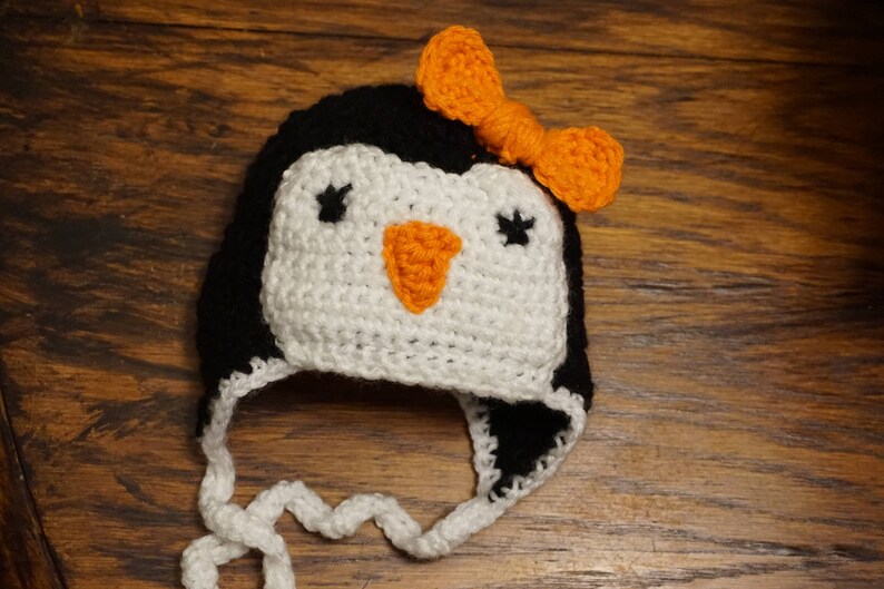 K&ouml;nnte beinhalten: Eine wei&szlig;e und schwarze geh&auml;kelte M&uuml;tze in Form eines Pinguins mit einer orangefarbenen Schleife. Die M&uuml;tze hat einen schwarzen Schnabel und schwarze Augen.