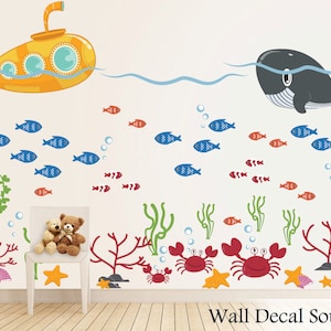 May include: A yellow submarine with portholes, a gray whale with a white belly, and red, blue, and orange fish swimming in a blue ocean.  There are also green seaweed, red coral, and orange starfish.  The text "Wall Decal Source" is at the bottom of the image.