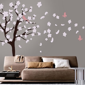 May include: A brown tree with pink flowers and a pink bird on a branch. The tree is on a gray wall with pink butterflies and flowers scattered around it. A brown couch with pillows is in the foreground.