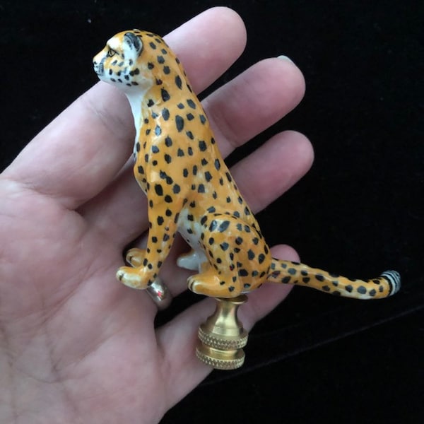Könnte beinhalten: Ein Lampenabschluss aus Keramik in Form eines sitzenden Geparden mit gelbem Fell und schwarzen Flecken. Die Gepardenfigur ist auf einem metallisch goldfarbenen Gewindesockel befestigt, der zur Befestigung eines Lampenschirms dient und gestuft ist.
