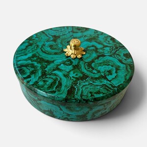 May include: A round lidded box featuring a vibrant green and black malachite pattern across its surface. The lid is topped with a small, ornate gold-toned metal knob that has a textured, leaf-like base and a circular, patterned top center.