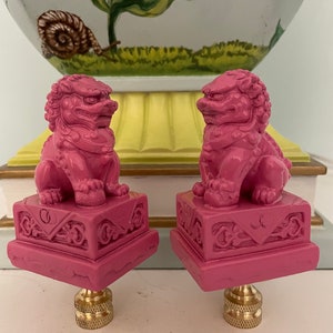 May include: Two pink ceramic foo dog lamp finials with gold accents. The foo dogs are sitting on square bases with intricate carvings.
