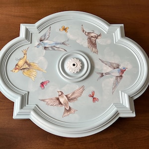 May include: A light blue, quatrefoil-shaped ceiling medallion with painted birds and butterflies. The center features a white, flower-shaped fixture. The birds are in flight, and the butterflies are in various colors. The medallion is a decorative element.