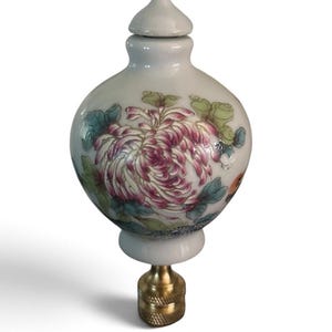 May include: A white ceramic lamp finial with a rounded, bulbous shape and a removable lid. The surface features a hand-painted floral design in shades of pink, green, and blue. It is attached to a textured, cylindrical brass-toned metal base.