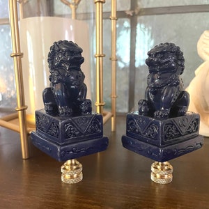 May include: Two blue ceramic foo dog finials with intricate details. Each finial is perched on a square base with a decorative design. The finials are made of a dark blue ceramic and have a glossy finish.