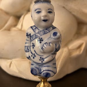 May include: A porcelain lamp finial featuring a small figure dressed in traditional clothing with blue and white floral patterns. The figure holds a small object and is mounted on a circular, textured brass base with a threaded metal connector.