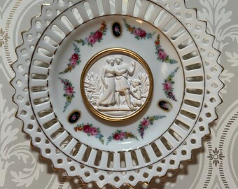 May include: A white porcelain decorative plate with a scalloped, pierced lattice rim featuring gold trim. The center displays a raised white relief of two figures in a landscape, surrounded by a gold border and a floral pattern with small black ovals.