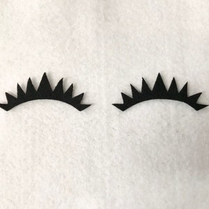 May include: Two black felt eyelashes with a jagged, pointed design. The eyelashes are on a white background.