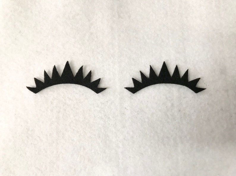 May include: Two black felt eyelashes with a jagged, pointed design. The eyelashes are on a white background.