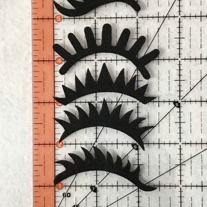 May include: Four black felt eye shapes with different lash designs. The shapes are arranged vertically on a gridded surface.