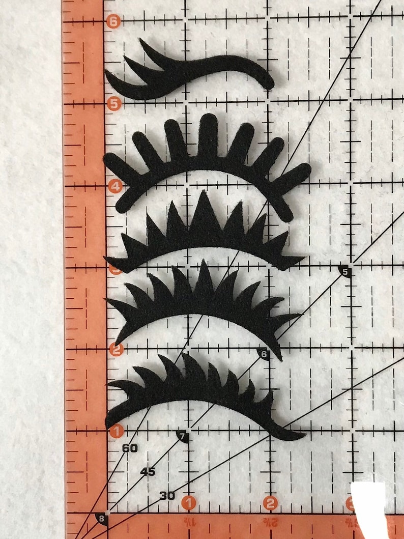 May include: Four black felt eye shapes with different lash designs. The shapes are arranged vertically on a gridded surface.