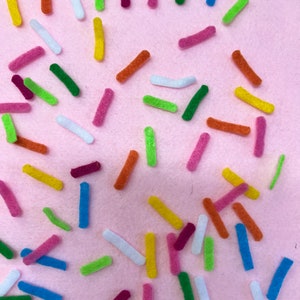 May include: A collection of colorful felt sprinkles in various shades of pink, green, blue, yellow, orange, and white. The sprinkles are scattered on a pink background.