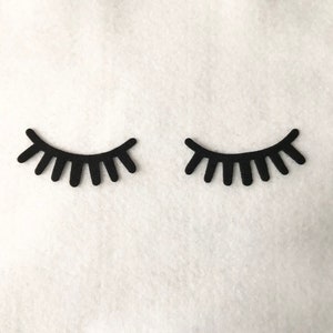 May include: Two black felt eyelashes, each with a curved shape and a row of individual lashes, are displayed against a white background.