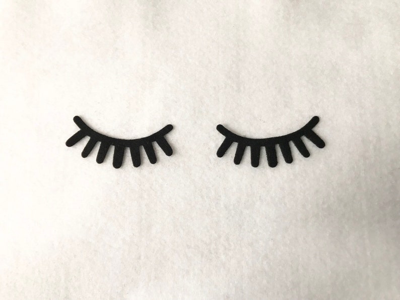 May include: Two black felt eyelashes, each with a curved shape and a row of individual lashes, are displayed against a white background.