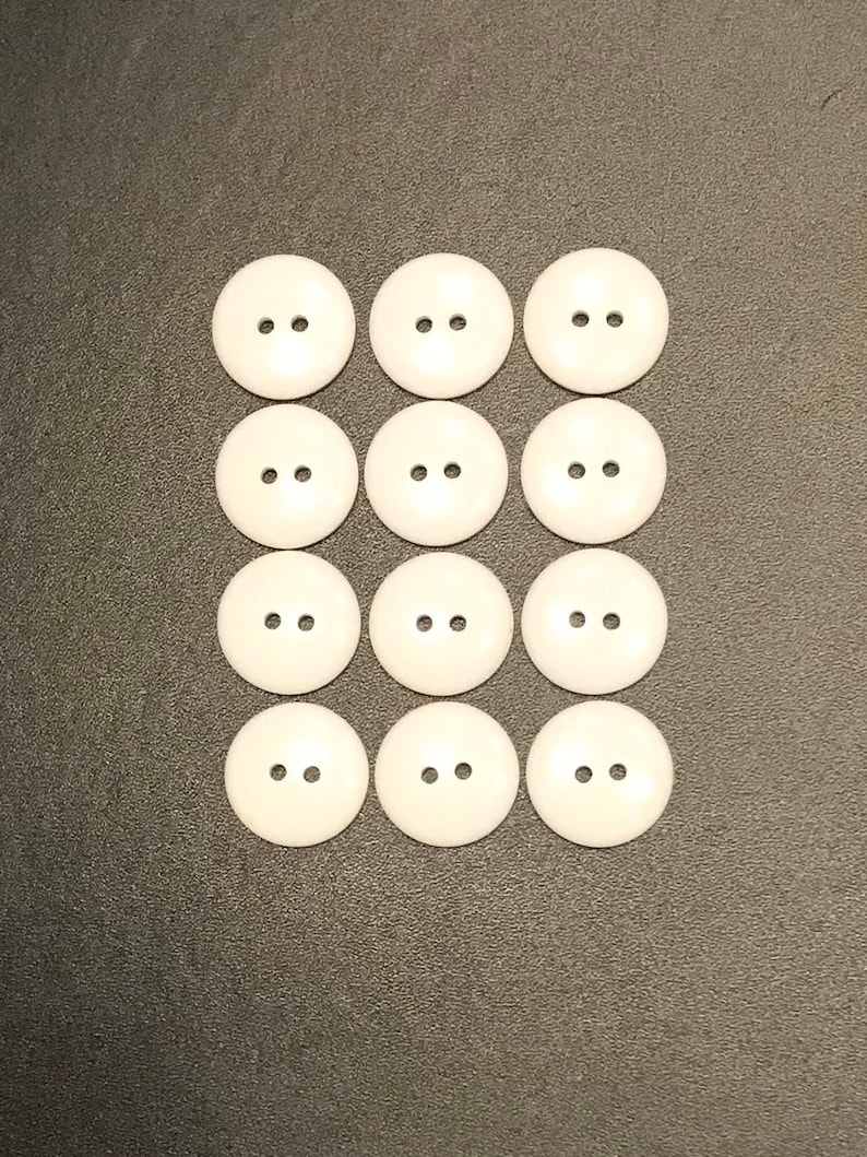 May include: Twelve white plastic buttons with two holes each. The buttons are arranged in three rows of four.