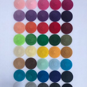May include: A collection of forty circular felt pieces arranged in an eight-by-five grid. Each piece features a concentric, stepped circular design with a raised center. The set includes a wide spectrum of colors, ranging from pastels to deep tones.