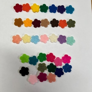May include: A collection of small, five-petaled flower shapes cut from felt fabric. The pieces are arranged in three horizontal rows and a scattered group below, featuring a wide variety of solid colors including red, orange, yellow, green, blue, purple, brown, white, black, and various shades of pink.