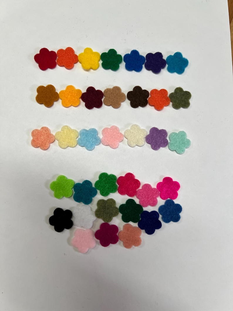 May include: A collection of small, five-petaled flower shapes cut from felt fabric. The pieces are arranged in three horizontal rows and a scattered group below, featuring a wide variety of solid colors including red, orange, yellow, green, blue, purple, brown, white, black, and various shades of pink.