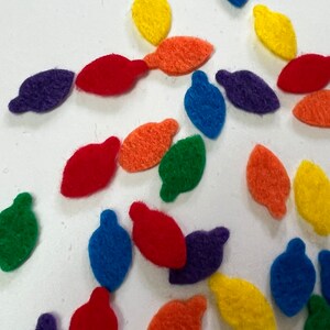 May include: Assortment of felt light bulb shapes in various colors, including red, orange, yellow, green, blue, and purple. The shapes are scattered on a white surface.