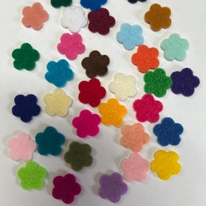 May include: A collection of small, five-petaled flower shapes made of felt in a variety of vibrant colors including red, blue, green, yellow, orange, pink, purple, white, black, and brown, scattered across a plain white surface.