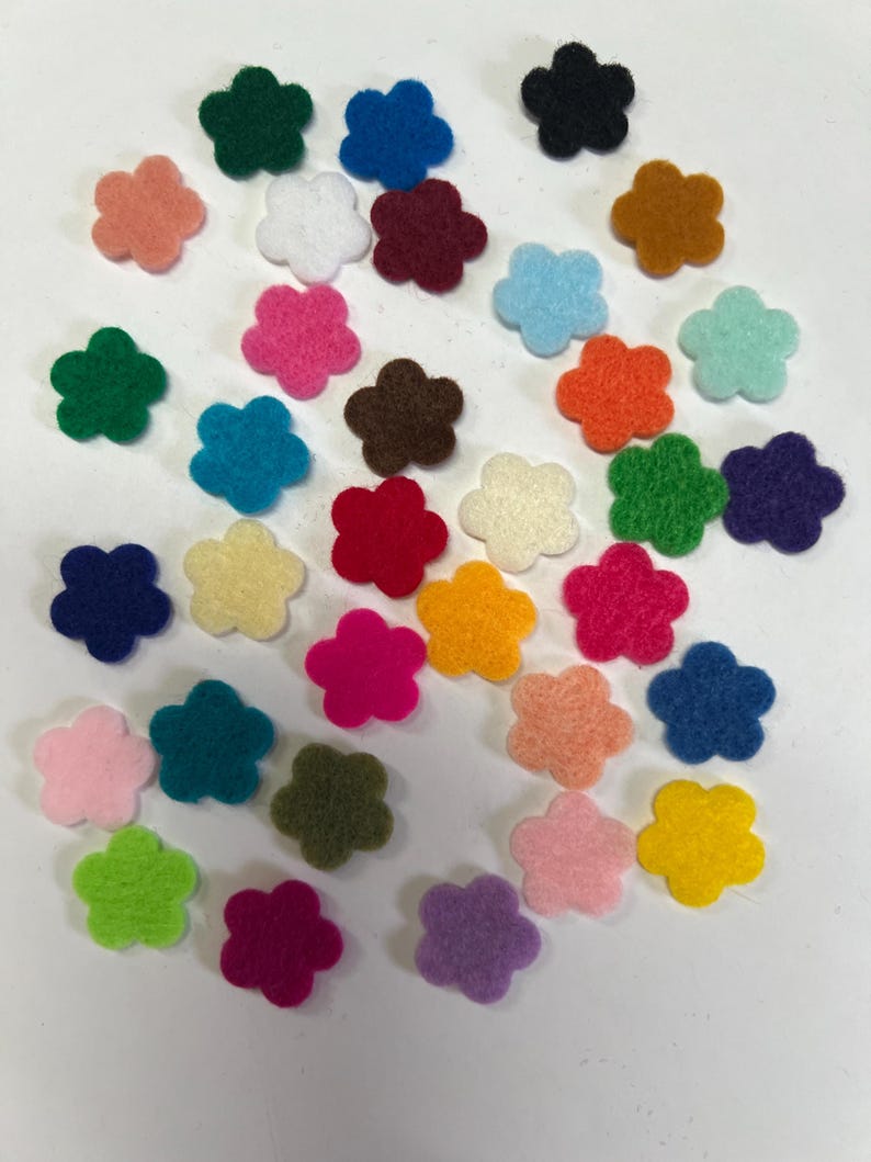 May include: A collection of small, five-petaled flower shapes made of felt in a variety of vibrant colors including red, blue, green, yellow, orange, pink, purple, white, black, and brown, scattered across a plain white surface.