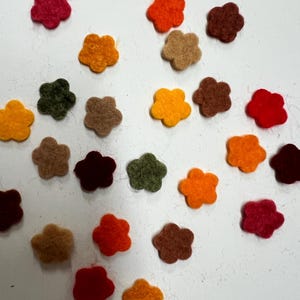 May include: Assortment of felt flower shapes in various colors, including red, orange, yellow, green, brown, and beige. The flowers are scattered on a white surface.