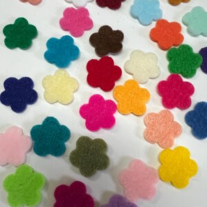 May include: A collection of small, flat, five-petaled flower shapes made of felt in a wide variety of vibrant colors including red, blue, green, yellow, orange, pink, purple, brown, white, and black, scattered across a plain white surface.