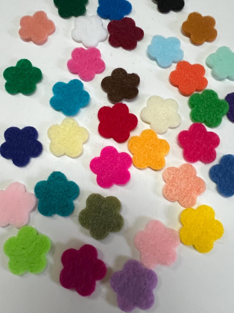 May include: A collection of small, flat, five-petaled flower shapes made of felt in a wide variety of vibrant colors including red, blue, green, yellow, orange, pink, purple, brown, white, and black, scattered across a plain white surface.