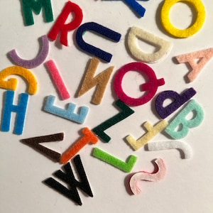 May include: A collection of felt letters in various colors, including red, blue, green, yellow, orange, pink, brown, and white. The letters are arranged in a random pattern on a white background.