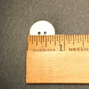 May include: A white plastic button with two holes, measuring approximately 1 inch in diameter. The button is placed next to a ruler for size reference.